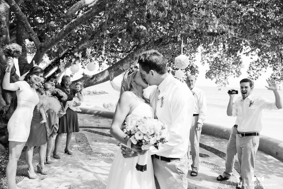 Black and white photo of couple kissing while their guests are cheering in the background.