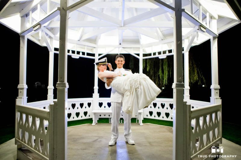 Military Groom picks up Bride for fun wedding couple portrait under white gazebo