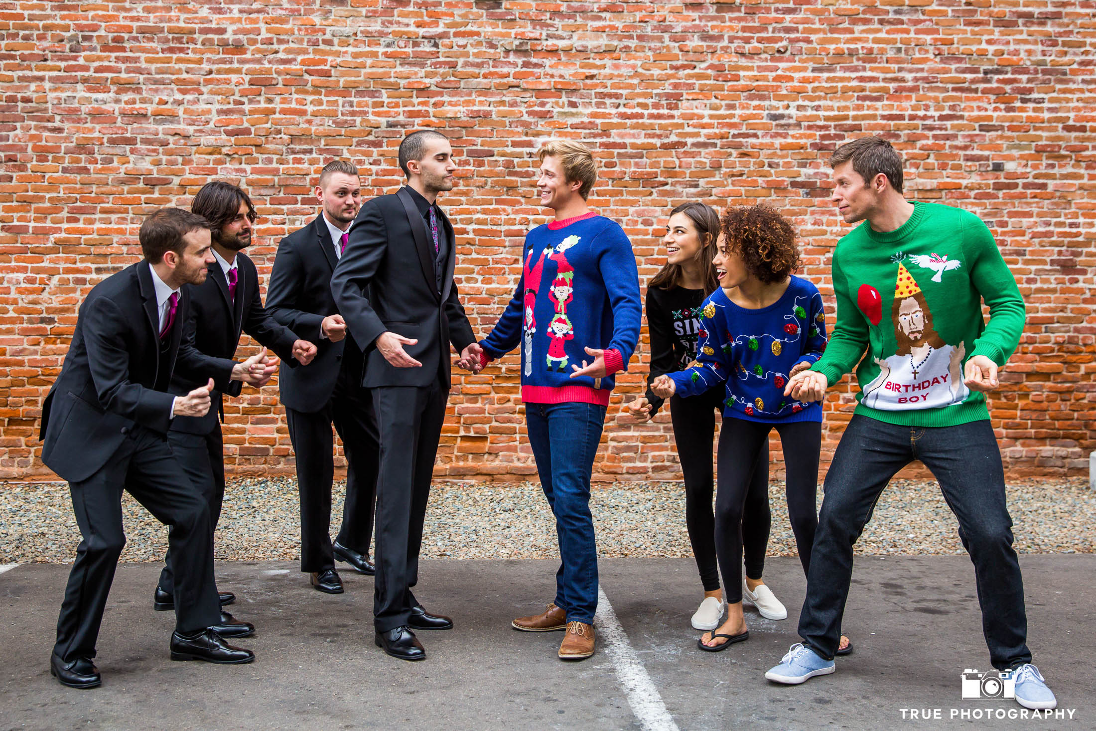 Groom and groomsmen snapping fingers in front of brick wall with Tipsy Elves in ugly christmas sweaters